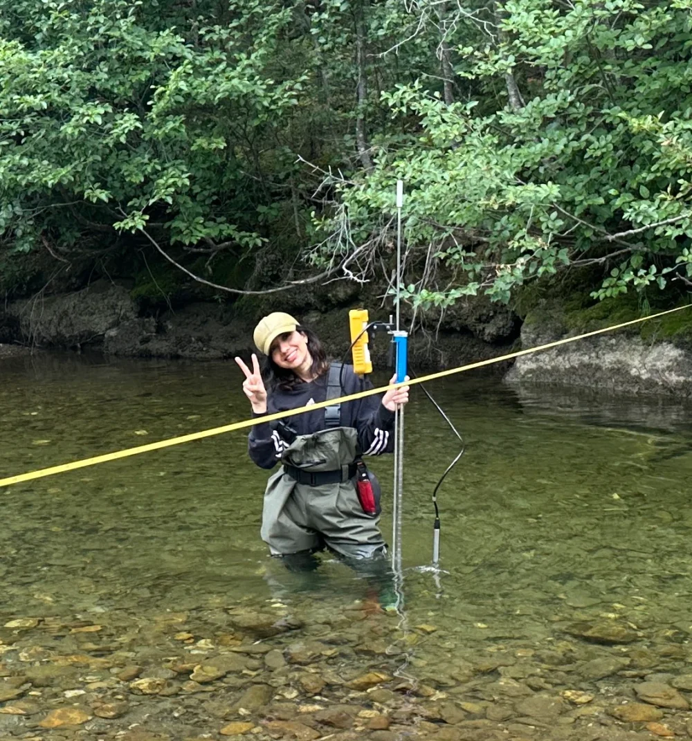 Behnoosh smiling in a stream with monitoring equipment and a measuring tape